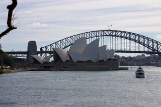  The image features a large white building with a distinctive arched structure, situated on a body of water. The building appears to be a bridge, with a boat passing underneath it. The scene is bathed in a soft, cloudy sky, creating a serene atmosphere. The boat is positioned towards the left side of the image, while the bridge spans across the entire width of the scene. The combination of the water, the bridge, and the boat creates a picturesque and tranquil setting.