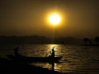  The image captures a serene scene of two people paddling a boat on a calm body of water, likely a lake, during sunset. The sun is setting in the background, casting a warm glow over the scene. The boat is positioned in the middle of the lake, with the two people sitting close to each other, enjoying their time on the water. The overall mood of the image is peaceful and tranquil, as the two individuals share a moment of relaxation and connection with nature.