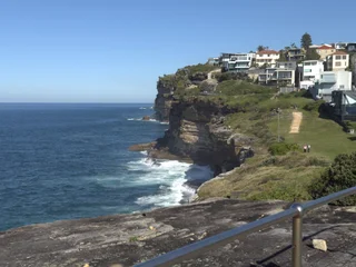 The image features a picturesque view of a cliff overlooking the ocean. The cliff is situated near a city, with a large building visible in the background. The ocean is calm, with a few small waves gently rolling in. The sky is a beautiful blue, creating a serene atmosphere. The scene is bustling with activity, as several people can be seen scattered throughout the area, likely enjoying the view and the surroundings. The combination of the cliff, ocean, city, and people creates a vibrant and dynamic landscape.