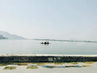  The image features a serene scene of a boat floating on a large body of water, possibly a lake or a river. The boat is situated in the middle of the water, and it appears to be a small, wooden boat. The water is calm and reflective, creating a peaceful atmosphere. The sky above the water is a clear blue, indicating a sunny day. The overall mood of the image is tranquil and inviting, with the boat gently gliding across the water, providing a sense of relaxation and escape from the hustle and bustle of daily life.