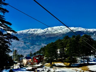  The image features a snowy mountain landscape with a ski lift running through the middle of it. The ski lift is carrying people up the mountain, likely to enjoy skiing or other winter activities. The scene is bustling with activity, as several people can be seen scattered throughout the area, likely preparing to ski or simply taking in the beautiful view. The mountain is covered in a thick layer of snow, creating a picturesque winter scene. The ski lift, people, and snowy mountain landscape all contribute to a lively and exciting atmosphere.
